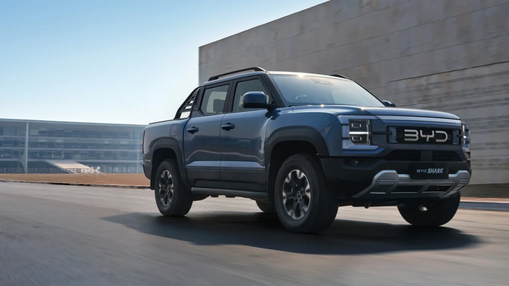 BYD Shark 06 plug-in hybrid pickup truck driving on city road, dual-cab PHEV pickup with rugged design and bold front grille